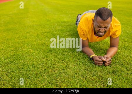 Portrait of a tired fit muscular afro american athlete sportsman doing plank exercise on a grass track field, young handsome male working hard doing Stock Photo