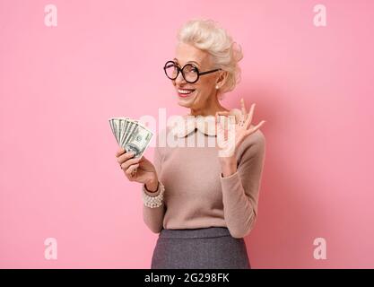 Smiling grandmother shows money cash and makes okay gesture, demonstrates that everything is fine. Photo of kind elderly woman wears eyeglasses on pin Stock Photo