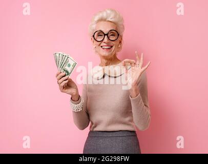 Smiling grandmother shows money cash and makes okay gesture, demonstrates that everything is fine. Photo of kind elderly woman wears eyeglasses on pin Stock Photo