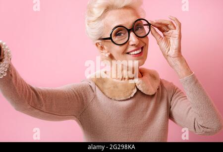 Cheerful grandma taking selfie on the phone. Photo of kind elderly woman wears eyeglasses on pink background. Stock Photo