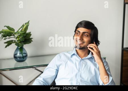 Portrait of smiling indian businessman in smart casual shirt talking on the smartphone, eastern man has pleasant phone conversation sitting in contemporary office, looks away and laughs Stock Photo