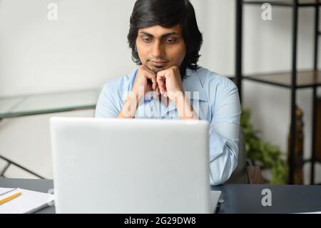 Focused clever indian man in smart casual shirt using laptop indoor, assembled hindu male office employee looks at the computer display, thoughtful guy working on a new project, researches the topic Stock Photo