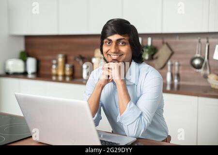 Happy indian freelancer man sits in the kitchen at home, looks at the camera and smiles cheerfully, confident successful hindu guy using laptop computer for outsourced work Stock Photo