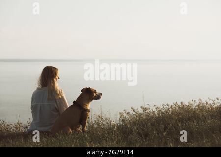 Woman and her dog sit in the sunset by the large lake or bay are Stock Photo