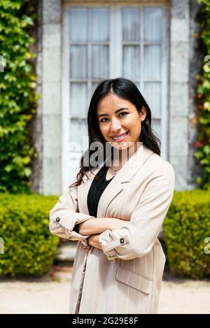 Confident woman in elegant outfit crossing arms standing against plant background Stock Photo