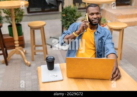 Headshot of young joyful African American businessman using phone and yellow laptop while working outdoors in the urban cafe, talking on mobile phone Stock Photo