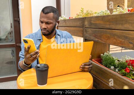 Busy pensive African American freelancer, student or businessman in casual clothing multitasking messaging on mobile phone, working on laptop drinking Stock Photo
