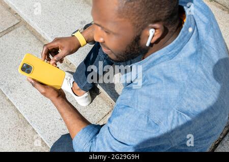 Back view of young African American man sitting on a park footway smiling and holding yellow mobile phone. Relaxed black guy in casual clothing having Stock Photo
