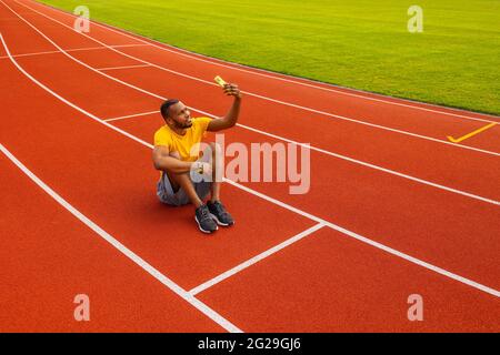 Handsome young afro american sportsman athlete runner is taking selfie at the stadium location. Smiling joyful black male laying on the field and Stock Photo