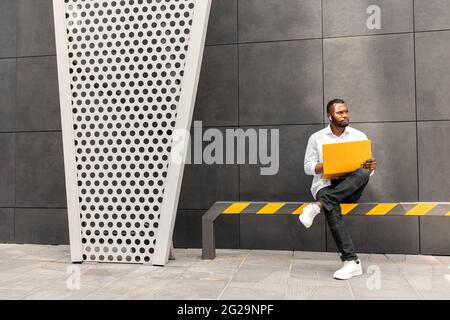 Serious young afro american man using yellow laptop computer while sitting on bench outdoors. Pensive businessman freelancer entrepreneur working Stock Photo