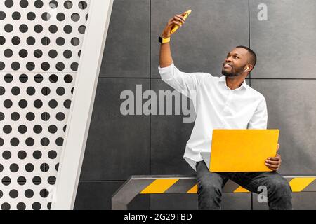 Handsome afro american man in trendy casual wear with yellow laptop in hand sitting in urban cafe outdoors, drinking take away coffee,hold remote talk Stock Photo