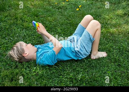 Barefoot child lying down the grass, plays with sensory fidget pop it toy. Flexible push bubble toy in hands of boy, playing outdoors at sunny day. Stock Photo