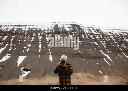 man in hat and checkered coat using vintage photo camera standing against snowy cliffs, Iceland Stock Photo