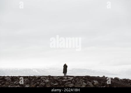 Back view of traveler standing on rocky terrain against misty mountains in snow, Iceland Stock Photo