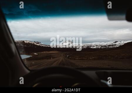 Magnificent view of countryside road and peak of snowy mountain through car windshield during trip through Iceland on cloudy day Stock Photo