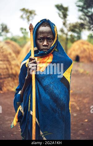 Teen boy holding wooden spear and wrapping in bright apparel while standing in middle of Mursi tribe village, Omo Valley, Ethiopia Stock Photo