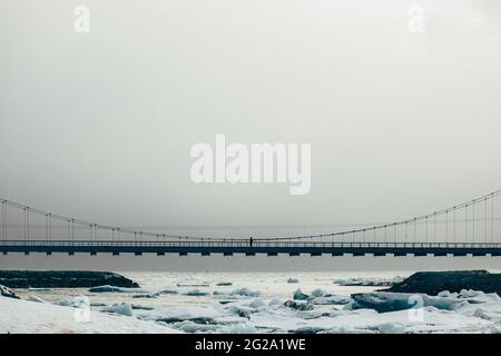 Unrecognizable person walking on iron bridge above white chunks of ice on calm lake water with remote snowy mountains and cloudy sky on background in Stock Photo