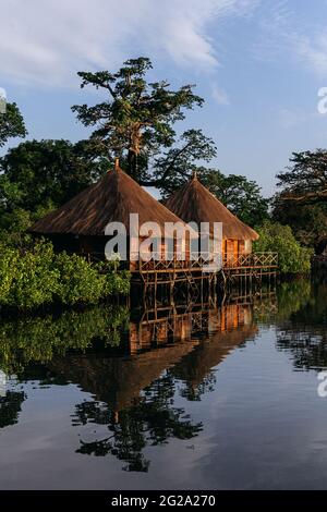 Wooden cottage with thatched roof and porch surrounded with trees by smooth sea over blue sky Stock Photo