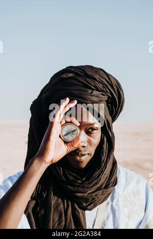 African male in keffiyeh smiling and holding vintage compass near eye while standing in Sahara Desert on sunny day Stock Photo