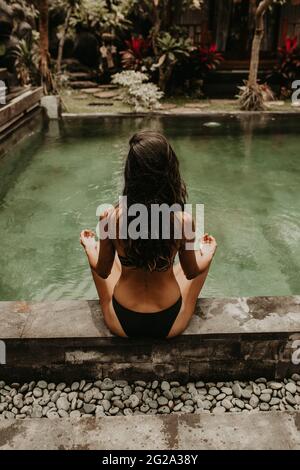 Back view of an anonymous ethnic female in black bikini doing yoga pose sitting and meditating alone near pool Stock Photo