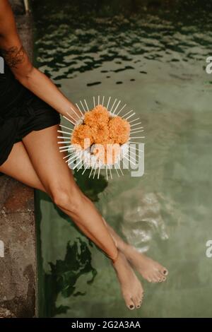 From above crop young slim female sitting on poolside holding composition of orange flowers Stock Photo