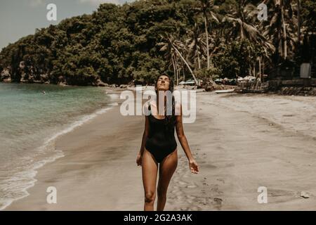 Slim young Woman in black swimsuit enjoying sun walking with closed eyes on sandy beach in sunlight Stock Photo