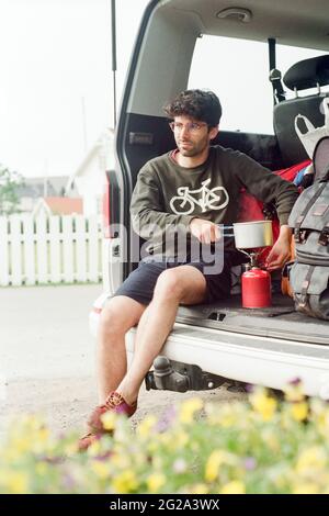 Young guy in eyeglasses heating pot on portable gas burner in trunk of automobile near road in Lapland Stock Photo