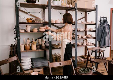 Clay made decoration in hands of anonymous woman in apron organizing shelves with finished products in pottery