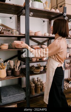 Clay made decoration in hands of anonymous woman in apron organizing shelves with finished products in pottery