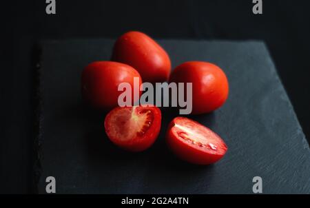 group of tomatoes, one cut in half, rests on a black board for serving Stock Photo