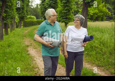 Happy senior couple with yoga mats walking along park path after sports workout in nature Stock Photo