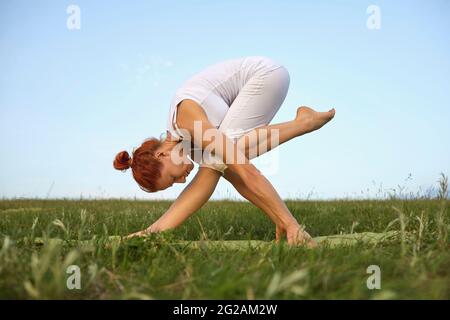 Young woman with flexible body practices yoga exercises on the grass against the sky. Stock Photo