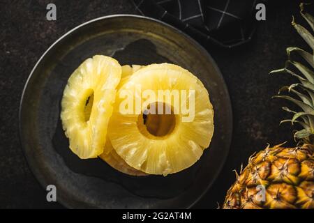 Canned sliced pineapple fruit on plate on black table. Top view. Stock Photo