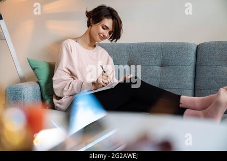 Lovely attractive young woman writing in a diary while relaxing on a couch at home Stock Photo
