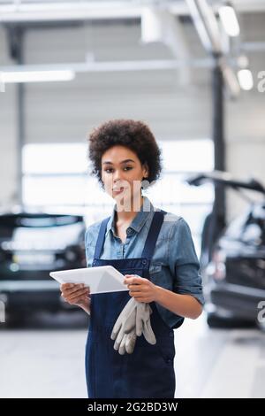 young african american mechanic holding digital tablet and looking at camera in garage Stock Photo
