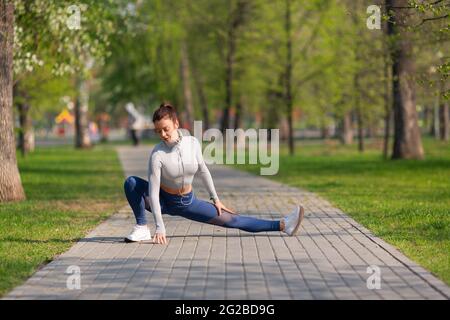 Full-length photo of a woman during stretchig workout in city park. She stretches the leg muscles and warms up before jogging workout.Sport and health Stock Photo