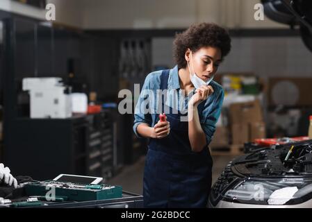 young african american mechanic holding protective mask with hand while working with motor of car in garage Stock Photo
