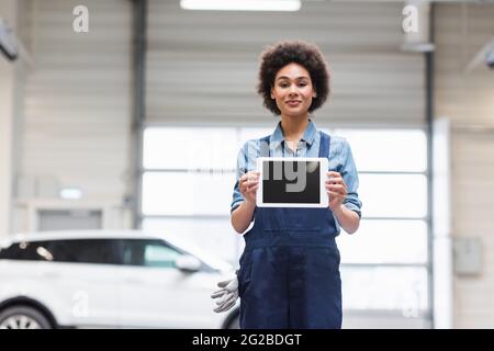 smiling young african american mechanic showing digital tablet with blank screen and looking at camera in garage Stock Photo