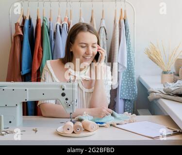 Happy seamstress talking on mobile phone while sitting at her workplace with sewing machine in studio Stock Photo
