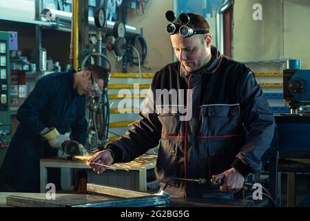 Welder working with autogenous welding tool in workshop while his colleague can be noticed in the back grinding. Stock Photo