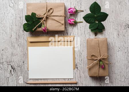 Gift boxes wrapped with kraft paper, pink rose buds and blank paper for text on gray background. Holidays concept. Mockup. Geometric flat lay Stock Photo