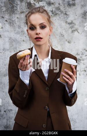 young woman in stylish outfit holding paper cup and doughnut against concrete wall Stock Photo
