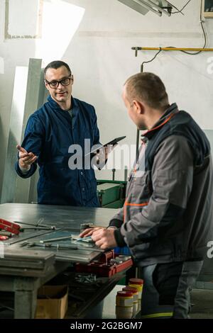 A tinsmith is measuring on a piece of the tin in his workshop and he's taking instruction from a colleague engineer who is holding tablet. Stock Photo