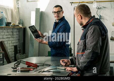 A tinsmith and his fellow colleague engineer are working together on a work task in their workshop, while the engineer is using tablet. Stock Photo