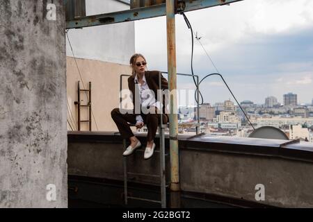 full length of young model in stylish sunglasses and trendy suit sitting near metallic stairs on rooftop Stock Photo