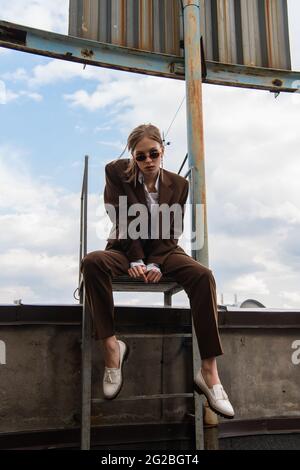 full length of young woman in sunglasses and trendy suit sitting on metallic rusty stairs on rooftop Stock Photo