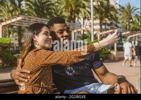 ALICANTE, SPAIN - Jun 09, 2021: Picture of Young Multiethnic Couple Sitting on a Bench Smiling Taking a Selfie with her Phone During the Holidays Stock Photo