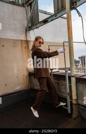 full length of young model in stylish sunglasses and trendy suit standing near metallic stairs on rooftop Stock Photo