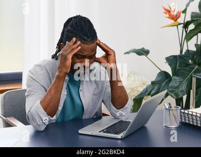 Remote work from home. African American man works at home workplace using laptop gets stress from distance work and lack of communication with people Stock Photo