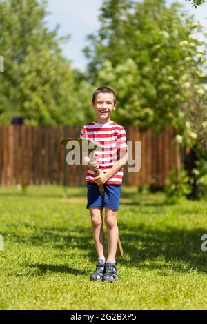 A little cheerful boy stands and sits on a shovel like a horse and jumps merrily, ready to work in the garden of a country house. Stock Photo